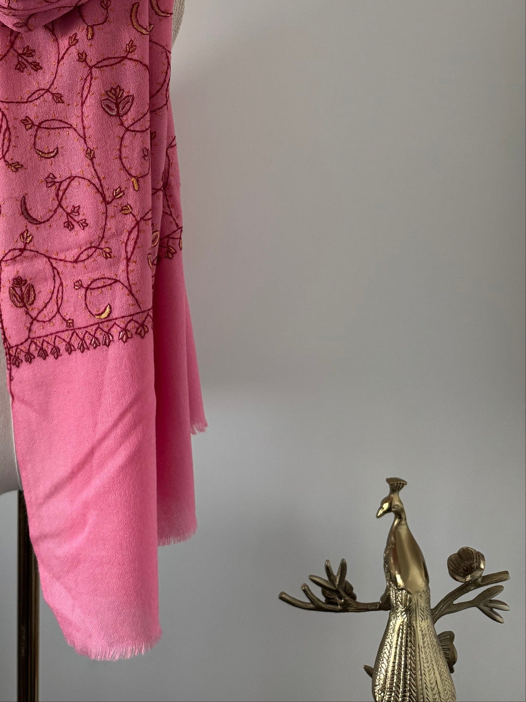 Pink scarf with embroidery next to a gold peacock figurine on a light gray background