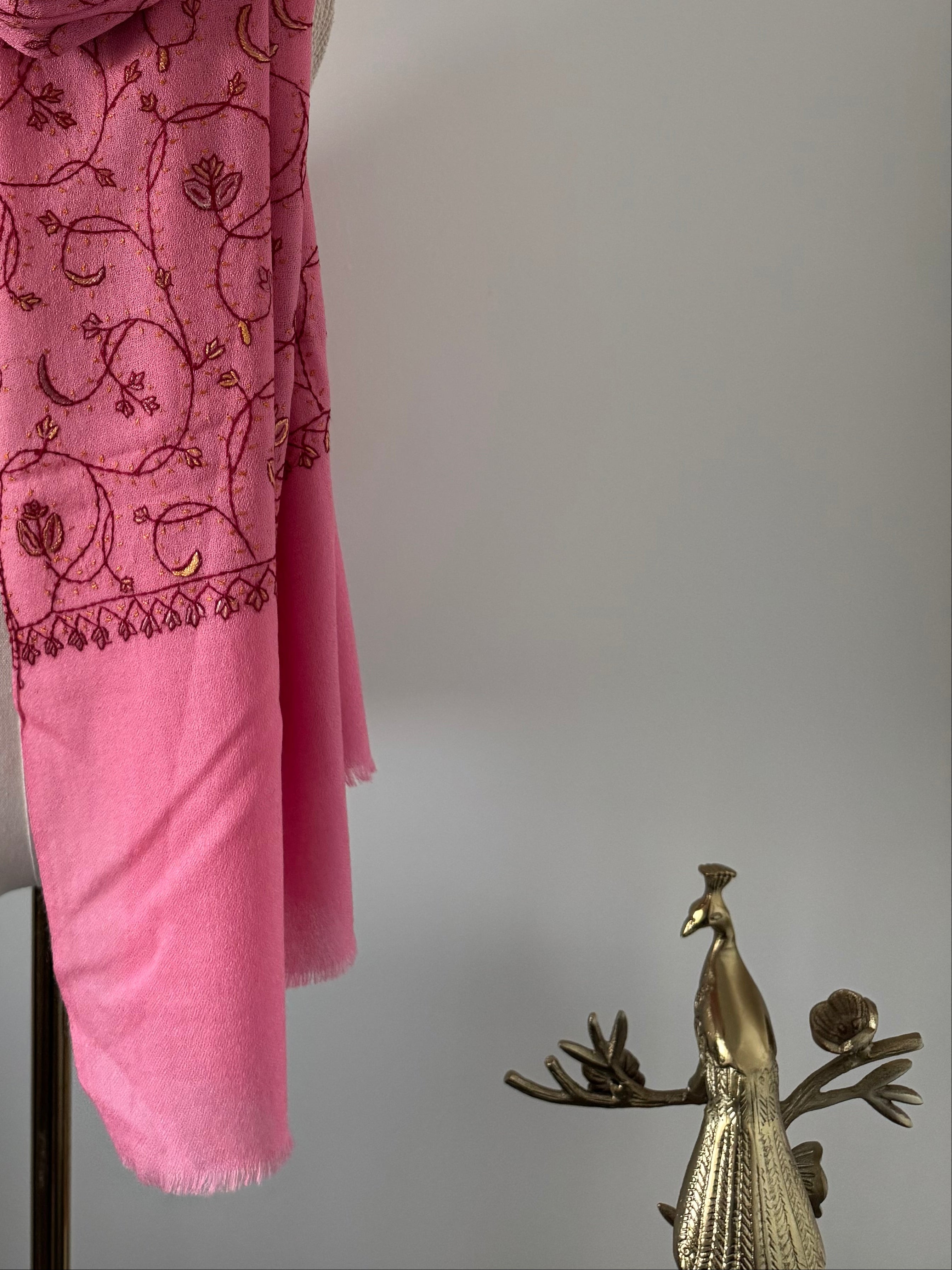 Pink scarf with embroidery next to a gold peacock figurine on a light gray background