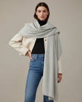 Woman wearing a gray scarf, beige jacket, black top, and blue jeans on a neutral background
