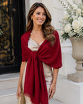 A woman wearing a deep burgundy shawl draped over a light beige satin dress, holding a small gold clutch, standing on white stone steps in front of a building entrance with a flower arrangement.