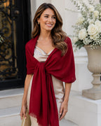 A woman wearing a deep burgundy shawl draped over a light beige satin dress, holding a small gold clutch, standing on white stone steps in front of a building entrance with a flower arrangement.