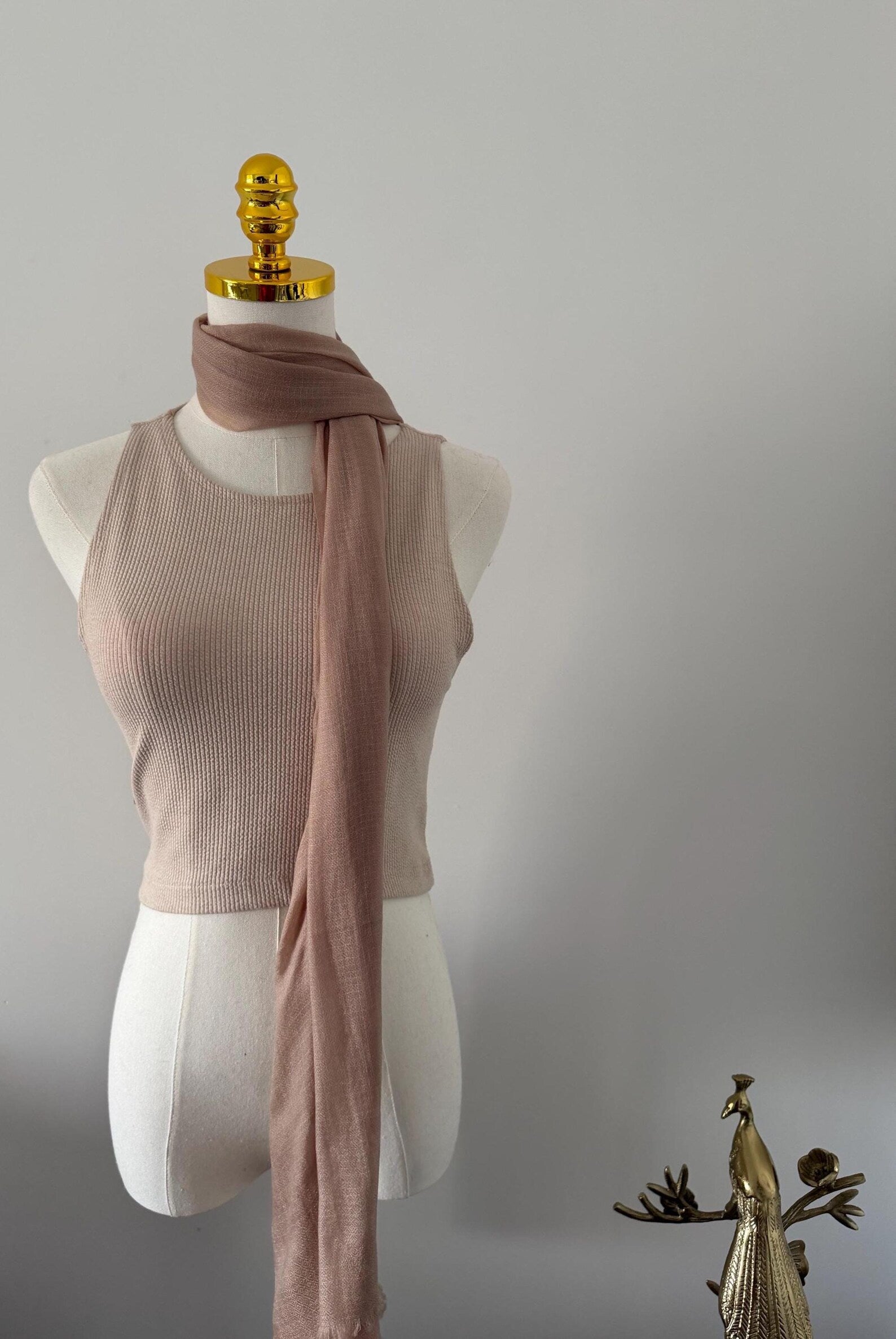 Mannequin wearing a beige ribbed tank top and pink scarf with a gold peacock ornament on a gray background