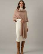 A woman wearing a beige shawl over a white outfit with brown boots on a plain background